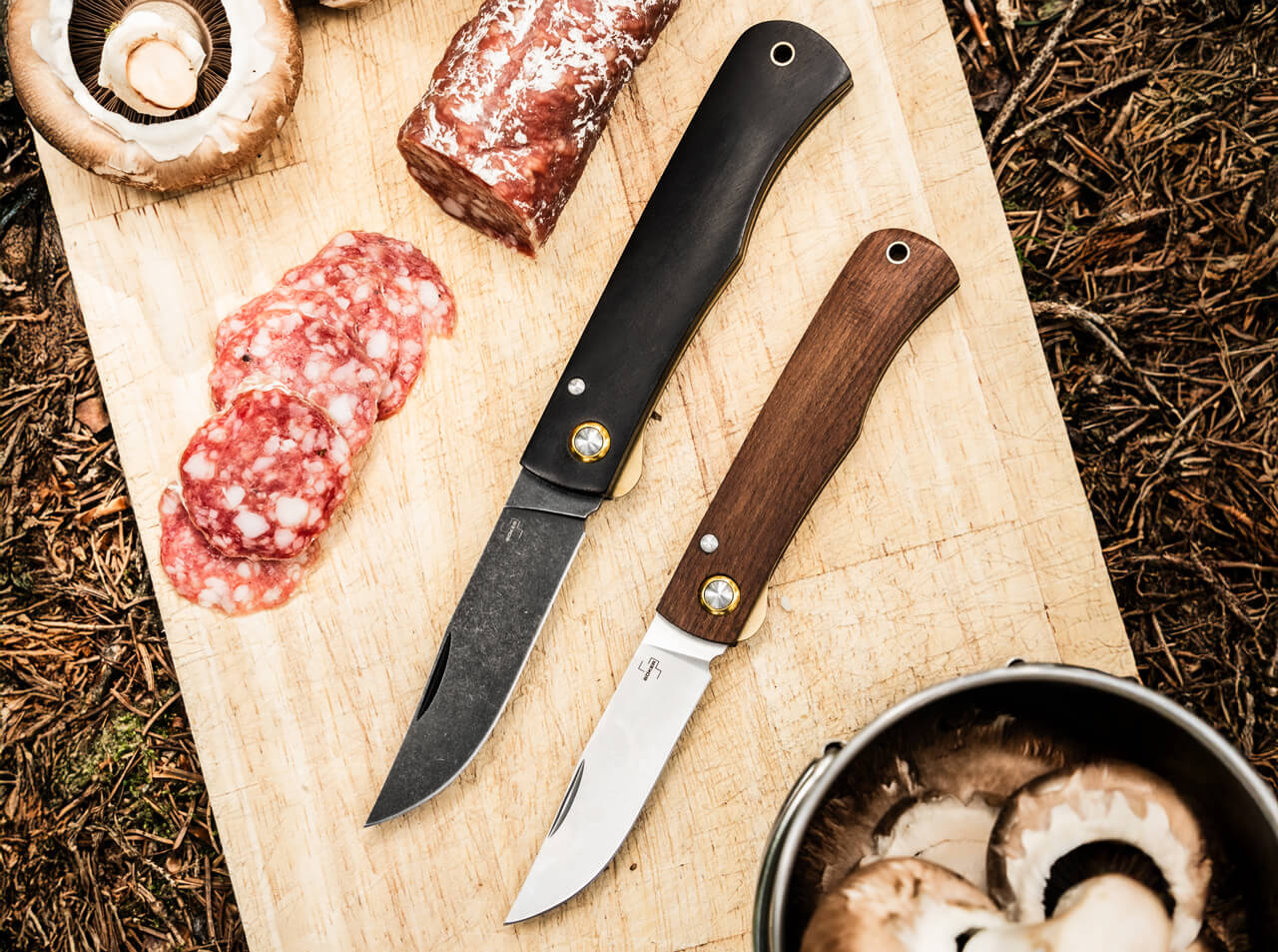 Two knives on a wooden cutting board with sliced salami and mushrooms.