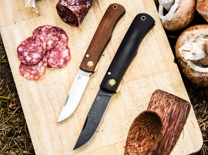 Two knives on a wooden cutting board with sliced salami and mushrooms.