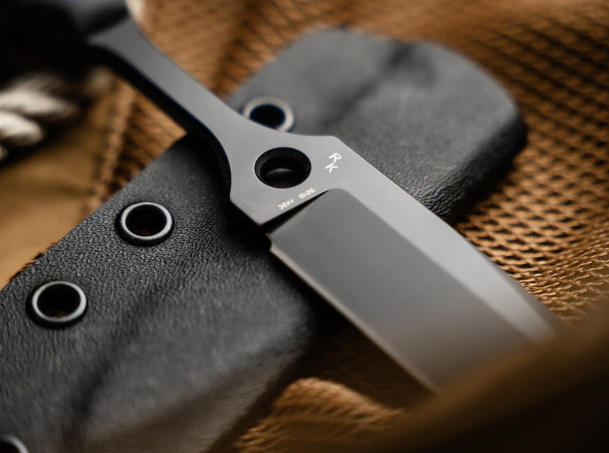 Close-up of a black metal knife with a matte finish resting on a black textured sheath over a brown woven fabric background.