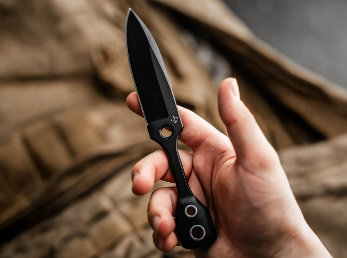 A hand holding a black tactical folding knife with a partially serrated blade, set against a blurred brown fabric background.