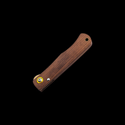 A folding pocket knife with a dark brown wood handle, shown diagonally against a solid black background. The handle features a circular lanyard hole near the top, a small silver rivet, and a yellow accent ring around a brass pin near the bottom.