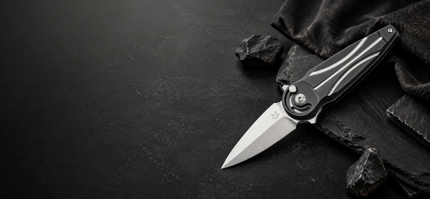 A folding pocket knife with a satin-finish silver blade and a black handle featuring white linear inlays, displayed diagonally on a dark textured surface with black slate chips and fabric.